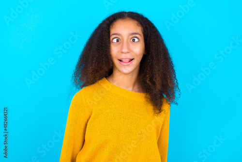 Funny beautiful teenager girl wearing yellow sweater standing against blue background makes grimace and crosses eyes plays fool has fun alone sticks out tongue.