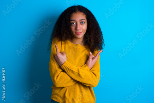 beautiful teenager girl wearing yellow sweater standing against blue background crosses arms and points at different sides hesitates between two items or variants. Needs help with decision