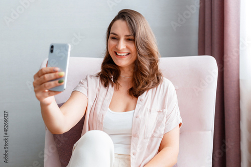 Beautiful tender young smiling caucasian 30s woman taking picture selfie using cell phone. Long haired brunette is photographing yourself on smartphone home interior.