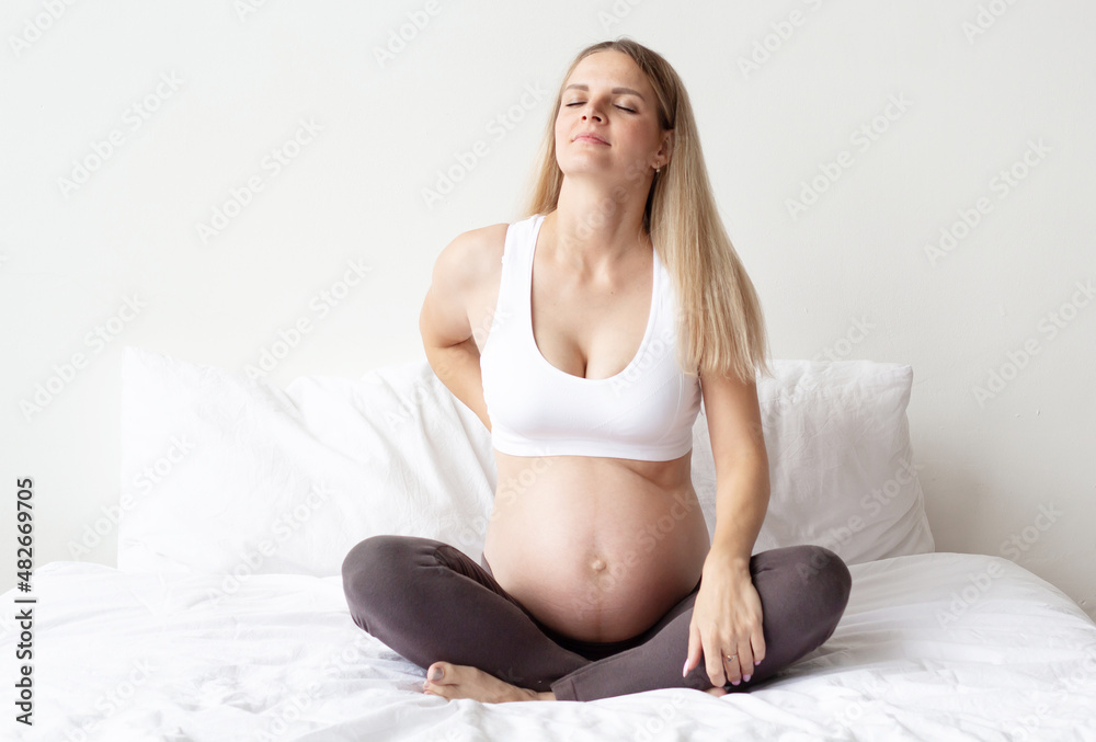 Beautiful blonde pregnant woman is sitting on the bed. Experiencing back pain. Problems bearing a child. Close-up. The background. Beautiful blonde pregnant woman is sitting on the bed. Experiencing back pain. Problems bearing a child. Close-up. The background.
