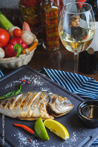 Grilled dorado fish on a plate with vegetables, lemon and a glass of white wine. 