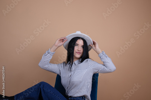 attractive brunette woman in blouse, hat and jeans on a blue chair on brown background. female fashion portrait. © OliaVesna