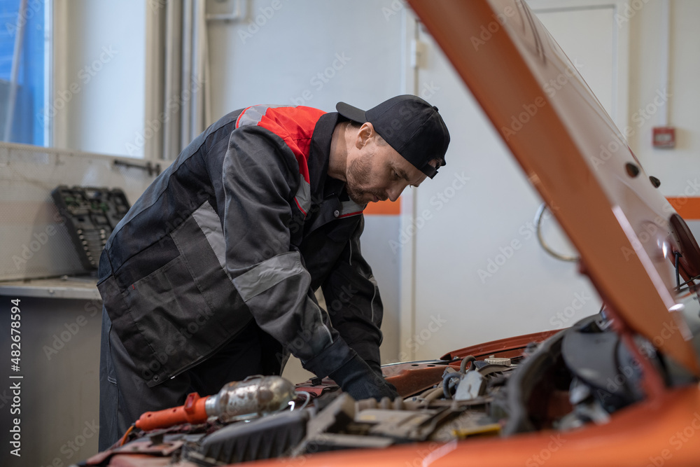 Young man in workwear bending over open car hood with engine while checking it before repairing in workshop Young man in workwear bending over open car hood with engine while checking it before repairing in workshop