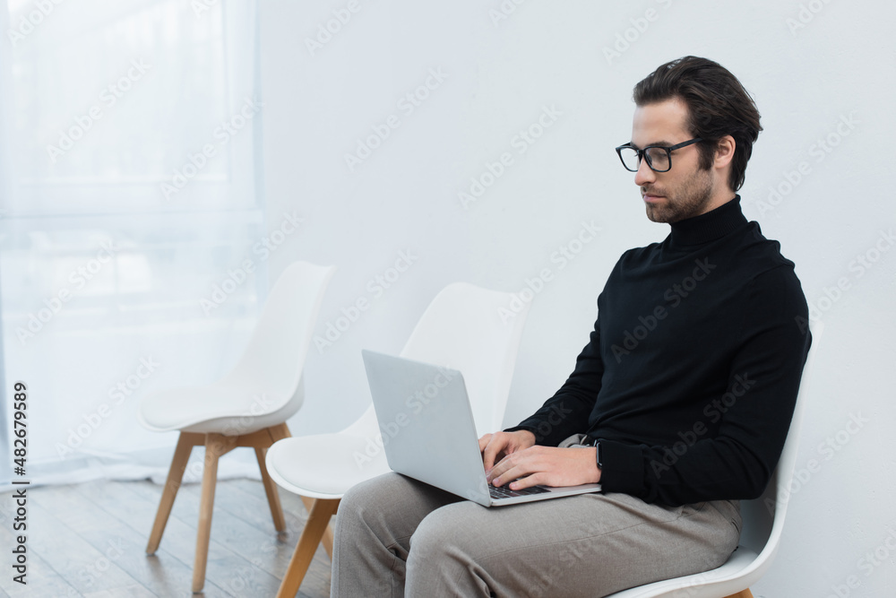 young freelancer in eyeglasses and black turtleneck sitting near grey wall and using laptop. young freelancer in eyeglasses and black turtleneck sitting near grey wall and using laptop.