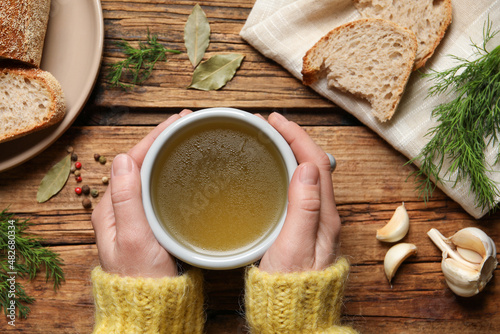 Woman with cup of hot delicious bouillon at wooden table, top view