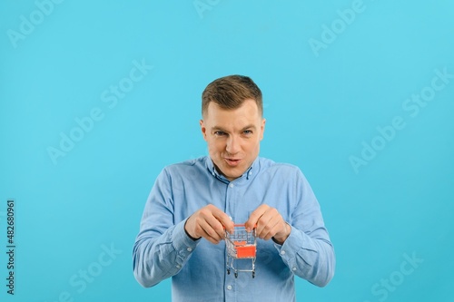 Young smiling man holding a mini shopping cart on a blue background. Sales concept. Place for text.