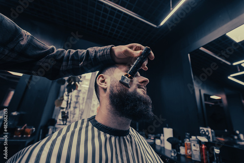 Barber works with a beard clipper. Hipster client getting haircut.