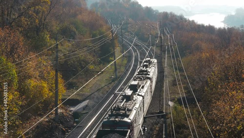 Freight train carries coal an electric locomotive by two-sided railway along the river and colorful forest at sunny autumn day. Aerial top view of locomative