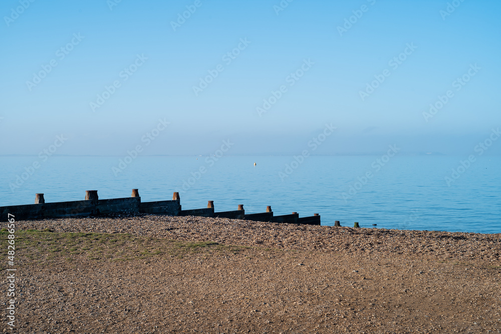 Wooden groyne water breakers on a pebble beach in Whitstable on a bright winter day with blue sky and blue sea. Isle of Sheppey can fairly be seen on the horizon. Wooden groyne water breakers on a pebble beach in Whitstable on a bright winter day with blue sky and blue sea. Isle of Sheppey can fairly be seen on the horizon.