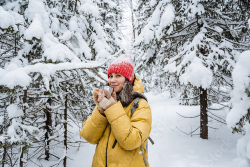 A girl holds a mug of hot tea in her hands. A woman on a walk in the winter forest drinks tea. Red hat on the head. Yellow jacket. Thermo glass in hand.