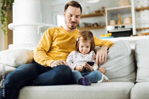 Single father and his small daughter using cell phone on sofa at home