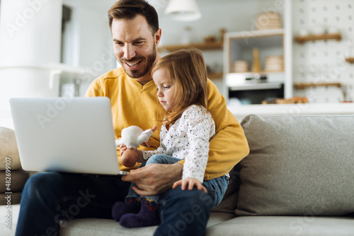 Young father and his small daughter using computer on sofa at home