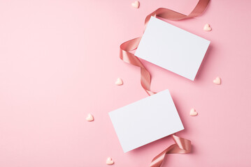 Top view photo of valentine's day decorations curly silk ribbon small hearts and two paper cards on isolated pastel pink background with copyspace