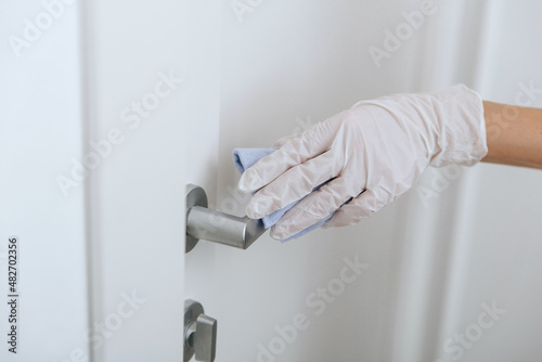 Cleaning door handle with blue wipe in white gloves. Sanitize surfaces prevention in hospital and public spaces against corona virus. Woman hand using towel for cleaning home room door link.