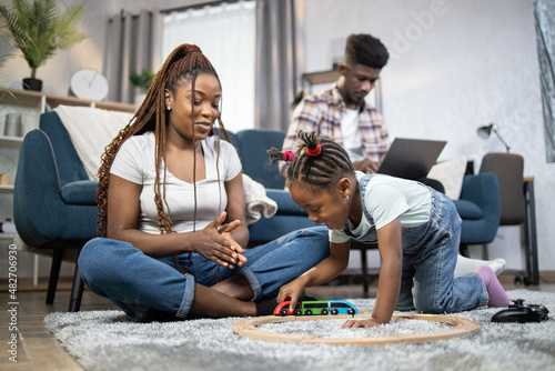 Joyful female child playing with happy mother on floor while father working on laptop. African american family spending time at cozy home.