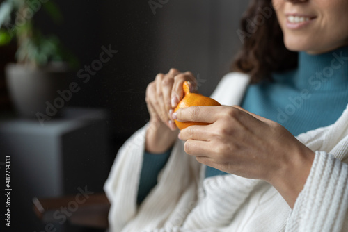 Closeup of smiling woman peeling off ripe tangerines. Joyful caucasian female enjoy fresh juicy citrus sitting at home relaxed covered with cozy blanket. Girl peel traditional winter fruit  soft focus