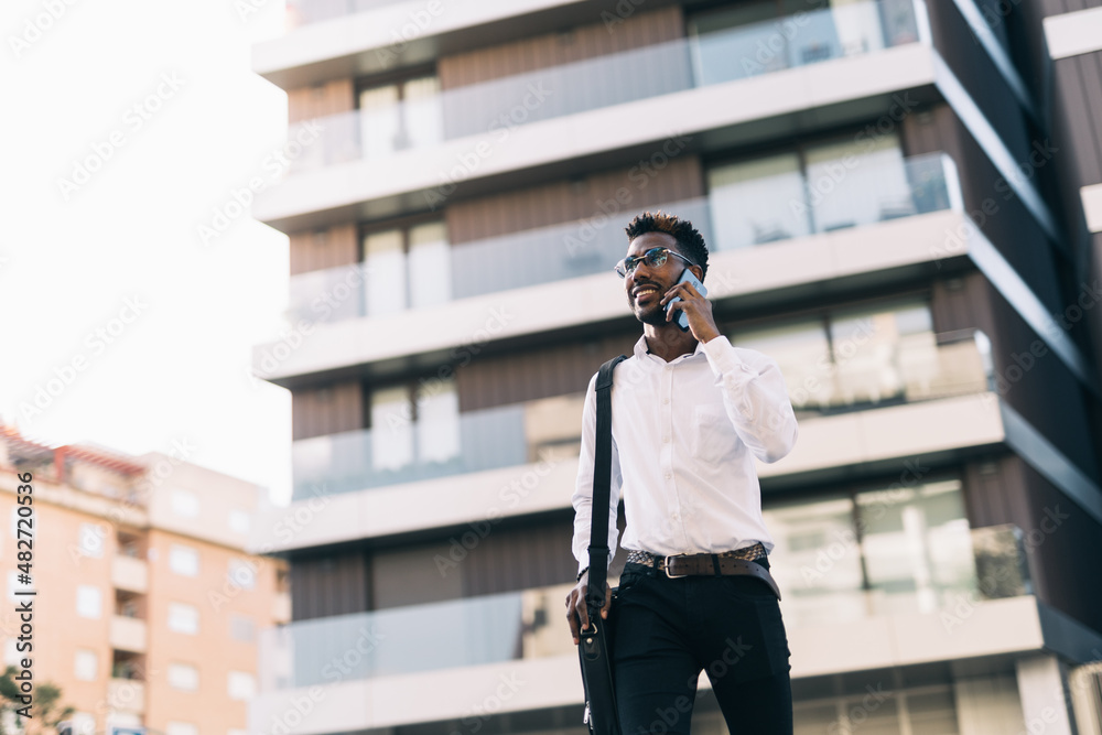 Fototapeta premium Happy and smiling young African American businessman walking across the street talking on his smart phone