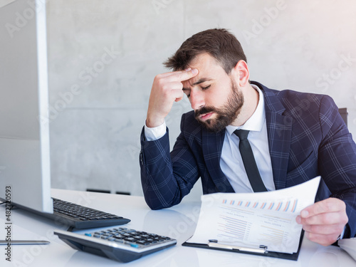 Modern male entrepreneur in suit with beard sitting at office desk, holding papers and looking worried.