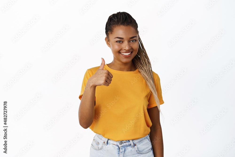Very good. Smiling satisfied Black girl shows thumbs up and nod in approval, like something, standing in yellow t-shirt over white background Very good. Smiling satisfied Black girl shows thumbs up and nod in approval, like something, standing in yellow t-shirt over white background