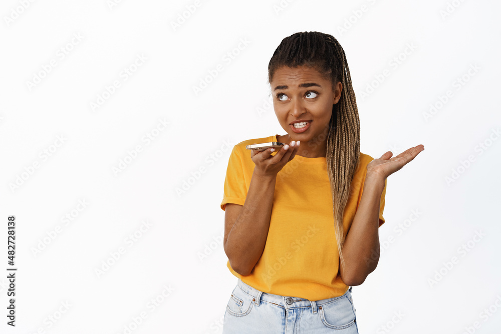 Image of black girl recording voice message, using voice app translator on mobile phone, talking on speakerphone, standing over white background Image of black girl recording voice message, using voice app translator on mobile phone, talking on speakerphone, standing over white background