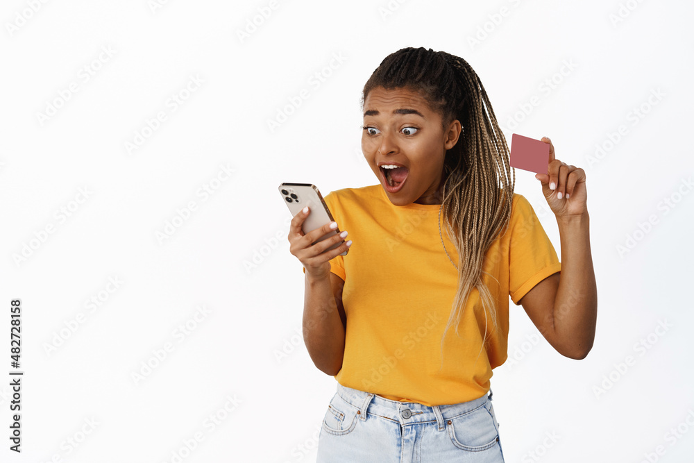 Excited girl looking at her mobile phone and showing credit card, amazed by banking app, standing over white background Excited girl looking at her mobile phone and showing credit card, amazed by banking app, standing over white background