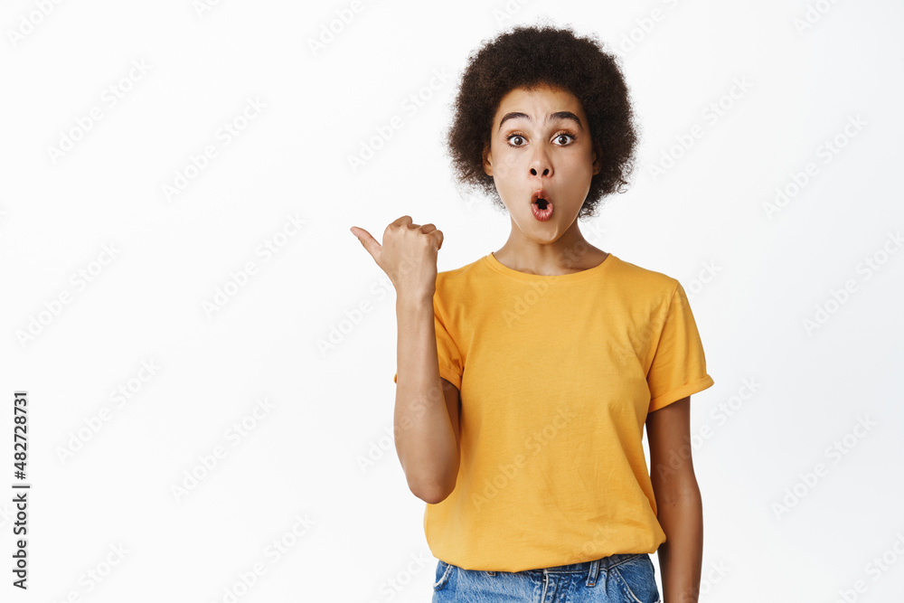Image of amazed black girl showing direction, pointing at empty space, company logo, standing against white background Image of amazed black girl showing direction, pointing at empty space, company logo, standing against white background