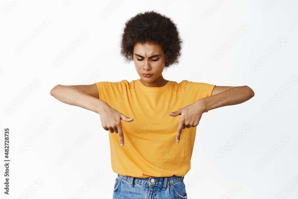 Young frowning black girl pointing fingers down and looking concerned, being doubtful about product, express disbelief or skepticism, white background Young frowning black girl pointing fingers down and looking concerned, being doubtful about product, express disbelief or skepticism, white background