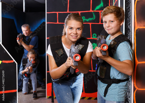 Cheerful teenager boy and his mother aiming laser guns at other players during laser tag game indoors