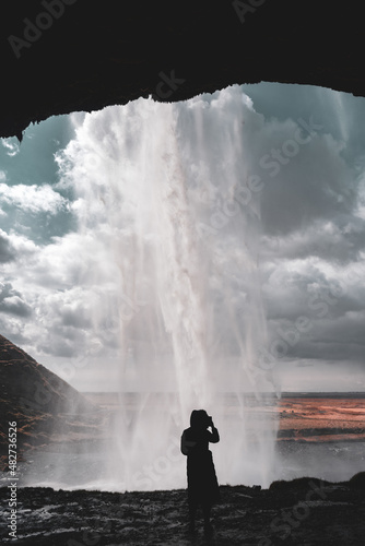 Vertical View of Seljalandsfoss Waterfall in Iceland from behind waterfall, with a woman silhouetted by the white cascade