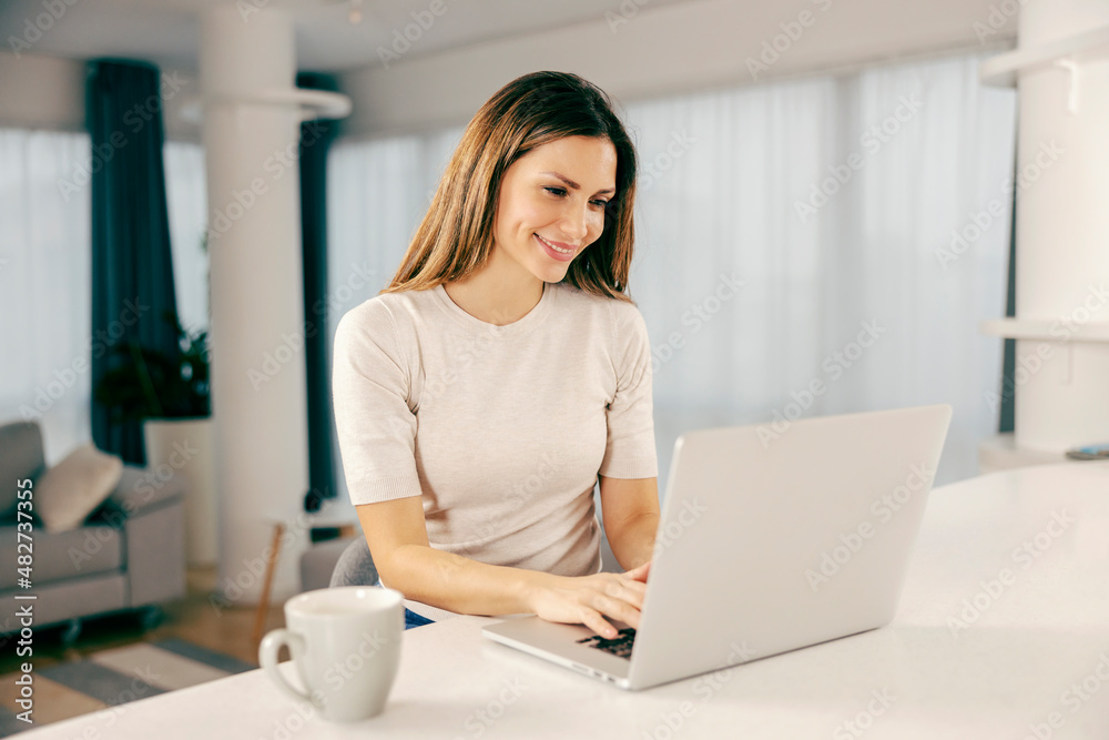 A happy woman using a laptop in kitchen at her home.