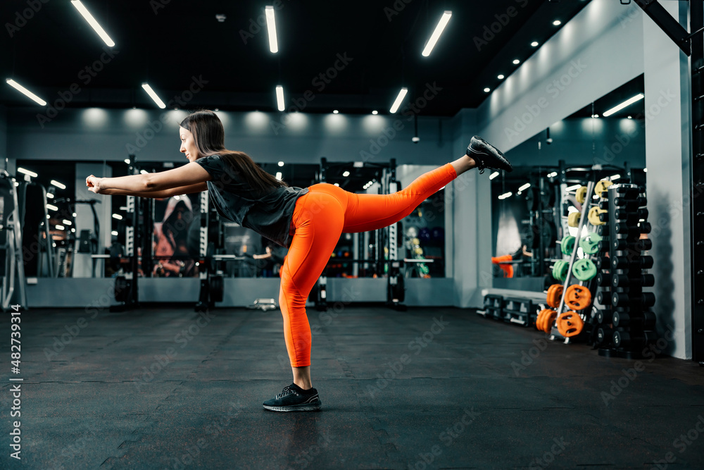 Fototapeta premium A fit sportswoman balances on one foot and does fitness exercises in a gym.