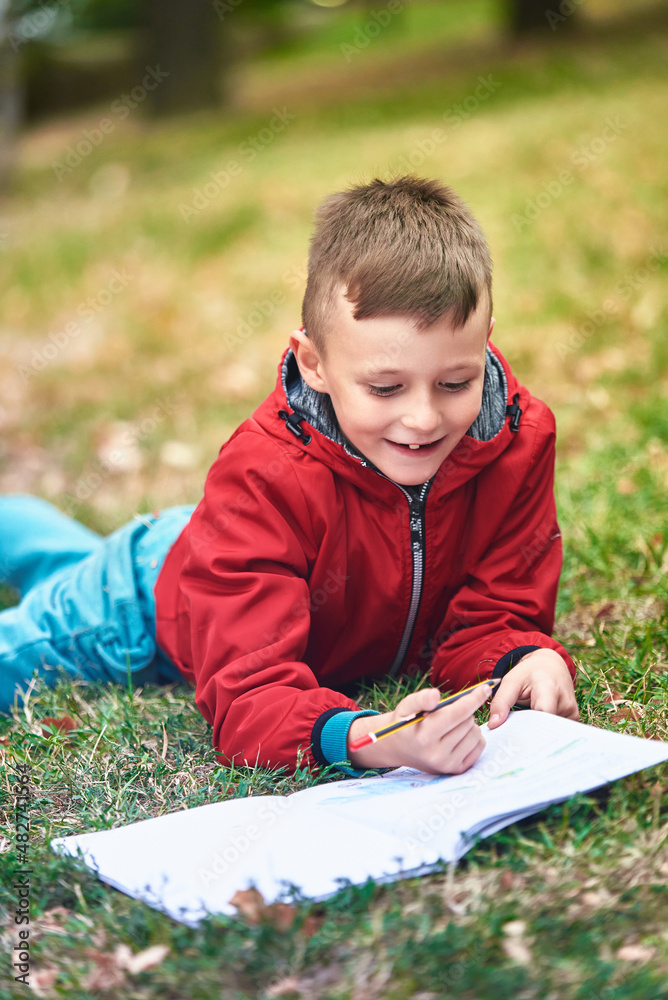 Cute young boy writing his homework in a park, lying on the grass, happy to learn and enjoying a nice sunny day in the park Cute young boy writing his homework in a park, lying on the grass, happy to learn and enjoying a nice sunny day in the park