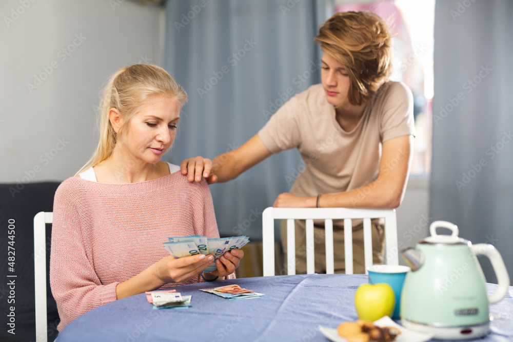 Portrait of woman giving money to son, chatting at table indoors