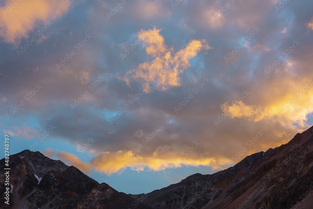 Scenic landscape with dark mountain range silhouette under evening blue sky with vivid orange sunset clouds. Sunlit orange cirrus clouds in sunset sky above mountains silhouettes at changeable weather