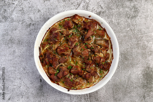 Braised pork with onions in a white baking dish on a dark grey background. Top view, flat lay