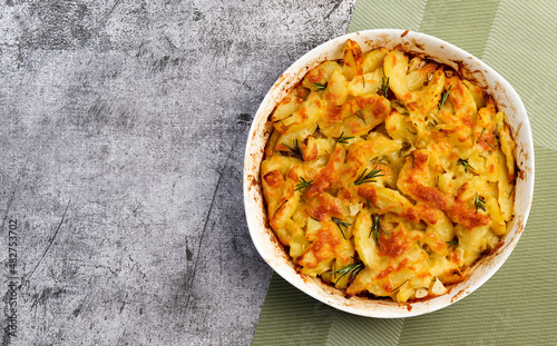 Potato casserole with cheese and rosemary in a white baking dish on a dark grey background. Top view, flat lay
