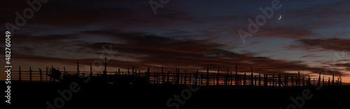 A panoramic image of a ranch corral silhouetted at dusk with some orange light lingering in the sky painting the clouds while the crescent moon hangs above the horizon to the right. photo