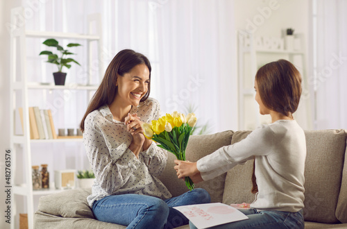 Loving daughter congratulates her mother on Mother's Day by giving her bouquet of flowers and handmade card. Happy young mother accepts gifts and greetings while sitting on sofa in living room.