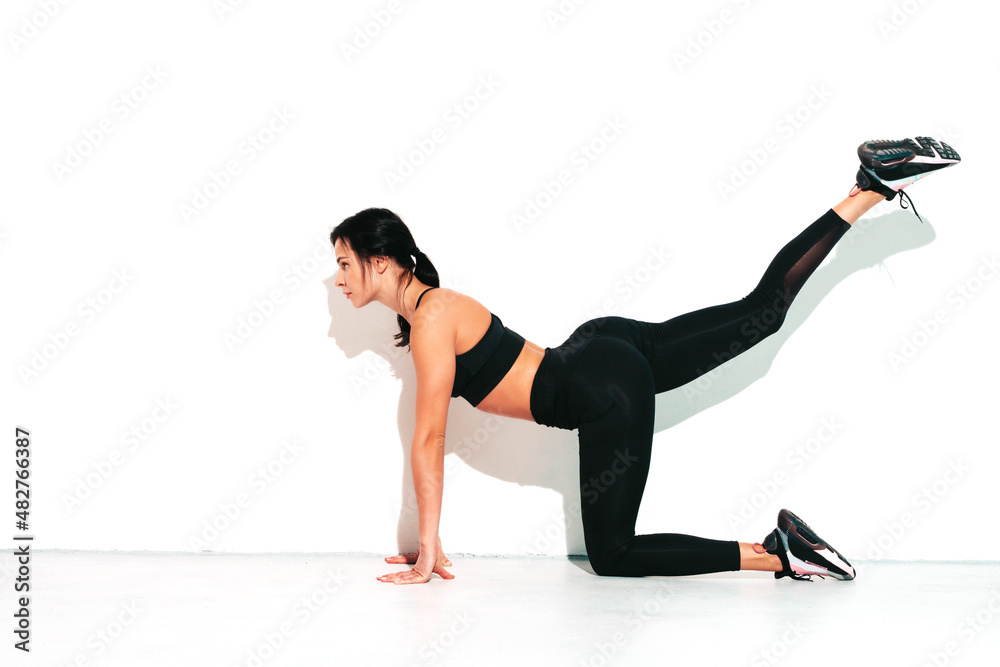 Portrait of fitness confident woman in black sports clothing. Sexy young beautiful model with perfect body. Female isolated on white wall in studio. Stretching out before training. Raising her leg Portrait of fitness confident woman in black sports clothing. Sexy young beautiful model with perfect body. Female isolated on white wall in studio. Stretching out before training. Raising her leg