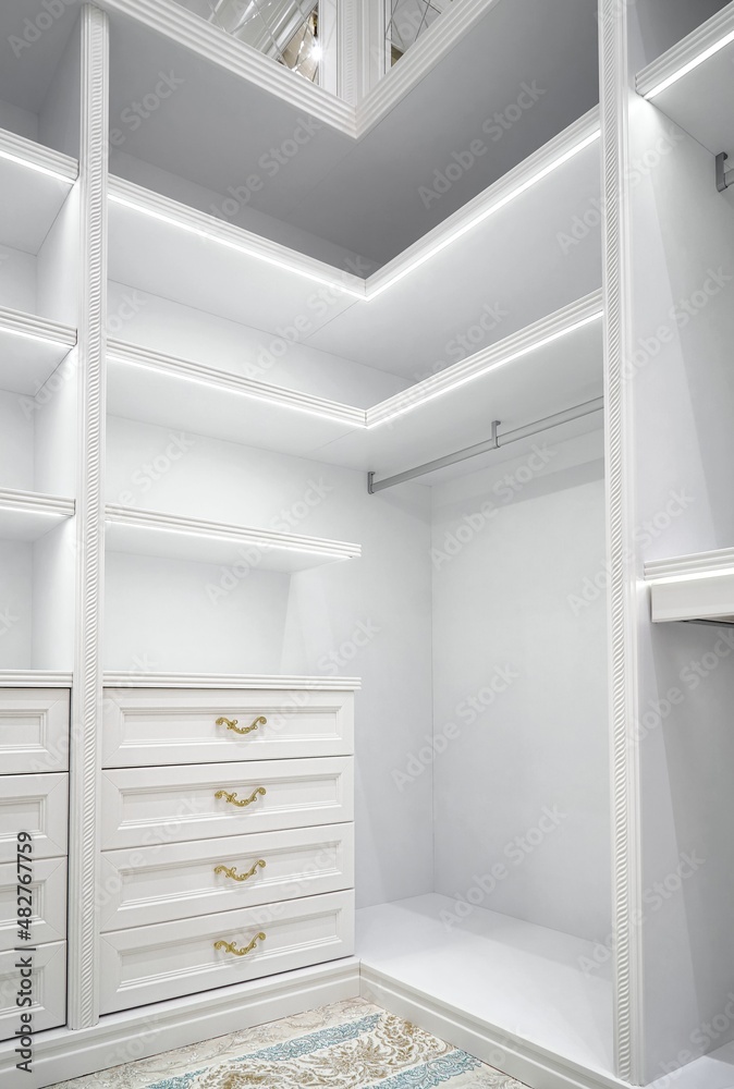 Fototapeta premium White corner wardrobe section with empty rack and led illumination on shelves in spacious walk-in closet in luxury apartment