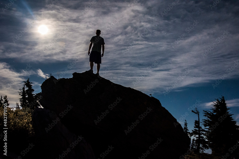 An athletic adventurous male hiker standing on a rock at night looking up at the stars.