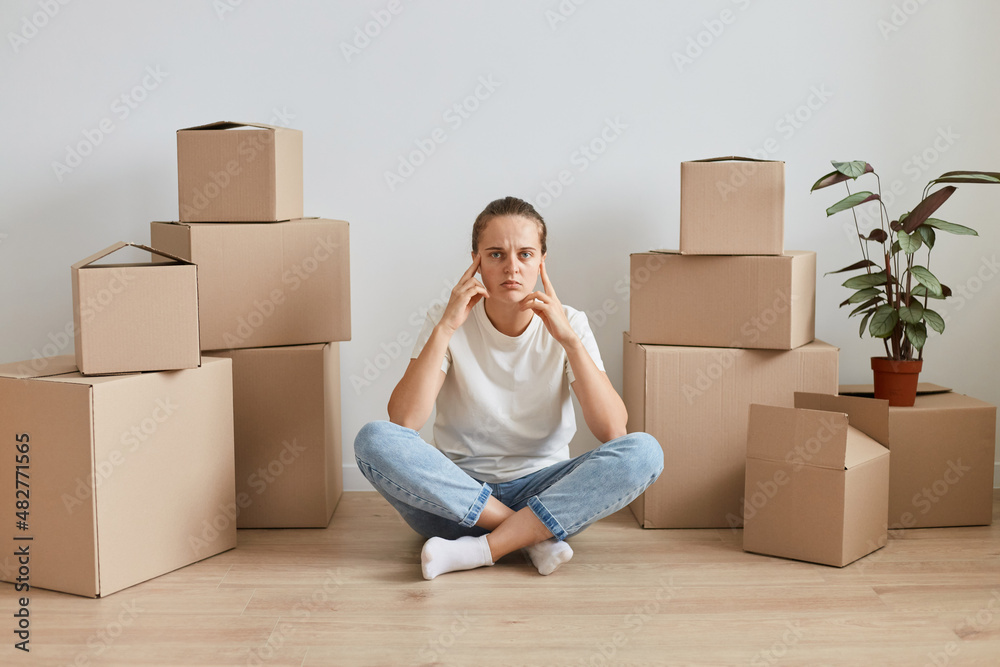 Indoor shot of tired pensive woman wearing white casual style t shirt, sitting on floor with crossed legs surrounded with cardboard packages, keeping hands on head, being thoughtful. Indoor shot of tired pensive woman wearing white casual style t shirt, sitting on floor with crossed legs surrounded with cardboard packages, keeping hands on head, being thoughtful.