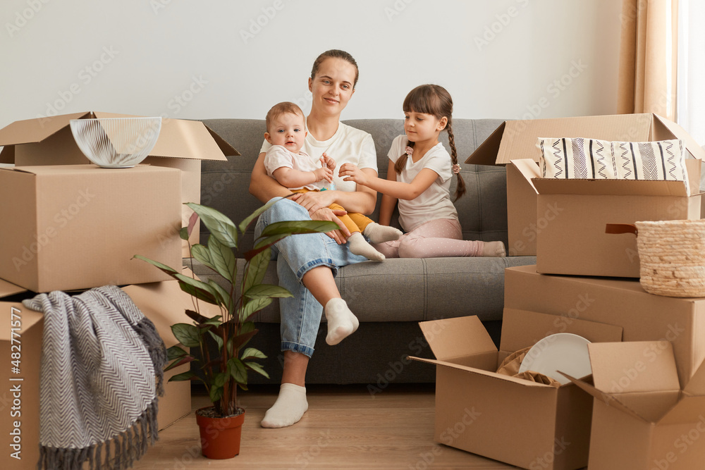 Portrait of happy family mother with daughters sitting on cough surrounded with cardboard boxes with personal piles, relocating to a new house, moving to renting apartment. Portrait of happy family mother with daughters sitting on cough surrounded with cardboard boxes with personal piles, relocating to a new house, moving to renting apartment.