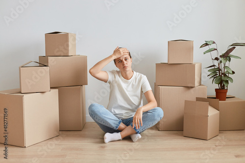 Portrait of exhausted woman wearing white casual style t shirt, sitting on floor with crossed legs surrounded with cardboard boxes with belongings, keeping hand on her head, looks tired.