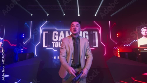 Excited adult announcer speaking to camera at start of professional esports competition on glowing stage with digital screen and neon illumination photo
