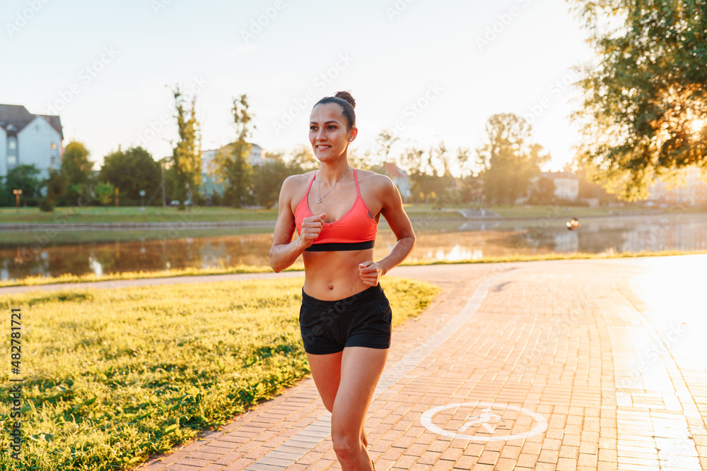 Full-length of young active sporty woman in sportswear running in city park on early sunny morning, sportswoman enjoying healthy lifestyle while jogging in nature. Sport, physical activity and health
