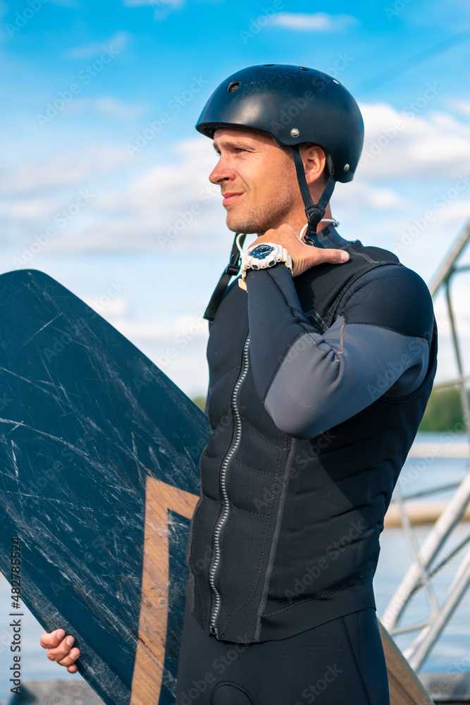 Focused wakeboarder in full gear standing on pier with board, intently looking away Focused wakeboarder in full gear standing on pier with board, intently looking away