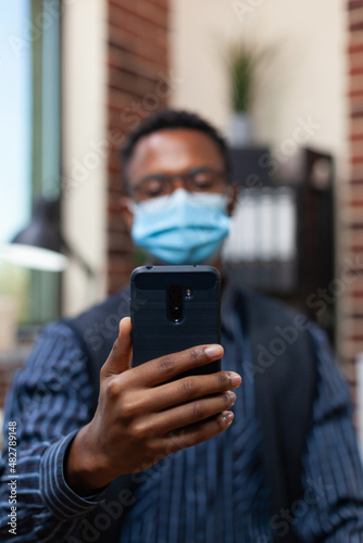 Closeup of entrepreneur holding smartphone in video call conference wearing covid protective mask and wireless earbuds. Detail view of startup employee smart phone in online interview. © DC Studio