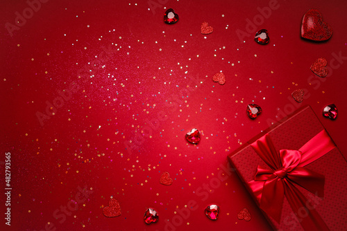 Valentine's Day gift in a red box tied with a red ribbon and red crystals, heart-shaped rubies, rhinestones, glitter on a red background with copy space.