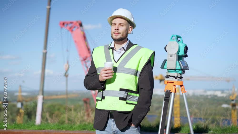 Medium shot portrait of young Caucasian man drinking coffee standing on construction site with tower crane at background. Handsome confident builder in uniform outdoors in sunshine looking away Medium shot portrait of young Caucasian man drinking coffee standing on construction site with tower crane at background. Handsome confident builder in uniform outdoors in sunshine looking away